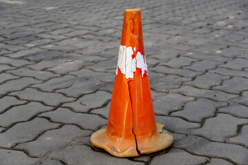 A cracked orange traffic cone stands alone on a cobblestone street, showing signs of wear and tear...