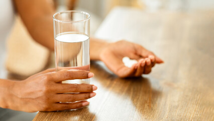 Unrecognizable black woman takes medicines with glass of water at home, cropped image, closeup