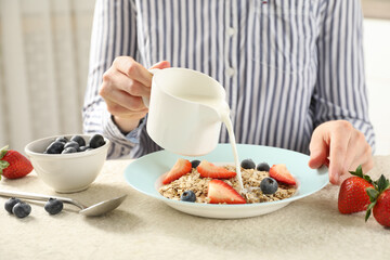 Woman pouring milk into bowl with oatmeal and berries at beige textured table, closeup