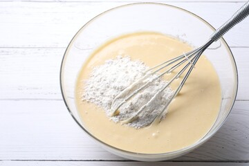 Whisk and bowl of dough on white wooden table, above view