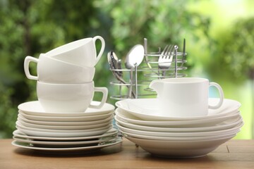 Different clean dishware, cups and cutlery wooden table against blurred green background
