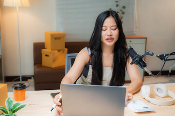 Young asian woman entrepreneur working at home using her laptop and recording a podcast, she is...