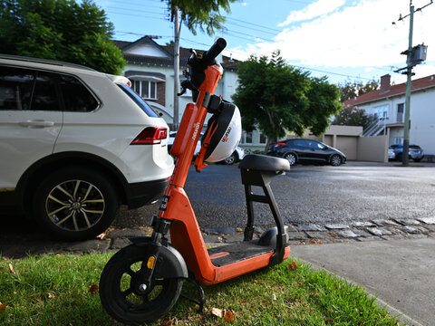Side view of an orange Neuron Mobility N4 e-scooter, parked on grass beside a suburban street during a sunny day