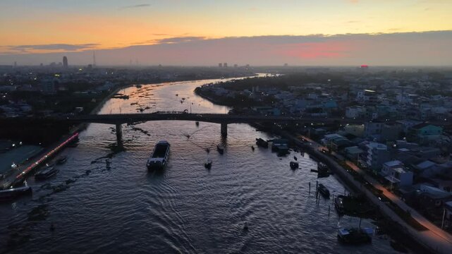 Drone view of a bustling Cain Tho river, Vietnam at sunrise, with various boats navigating the water, 
vehicles move steadily across Cai Rang bridge.
