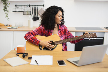 Young adult woman with curly hair playing guitar, watching online tutorial on laptop in kitchen. Home learning environment, creative self-improvement, indoor activity emphasizing personal development.