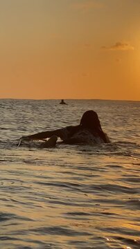 Long-haired young man surfer swims on a surfboard in the ocean at sunset.Vertical video