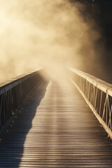 Obraz premium A wooden pedestrian bridge enveloped in dense fog, creating a mysterious and serene atmosphere as the light filters through in a golden hue.