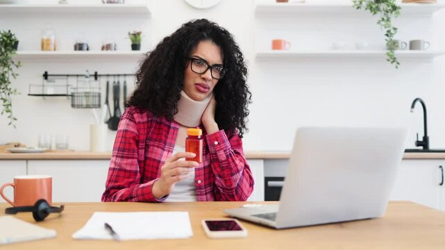 Young African woman in neck brace consulting online, looking at medication bottle, seated indoors, wearing red flannel, expressing discomfort, suggesting treatment or injury.