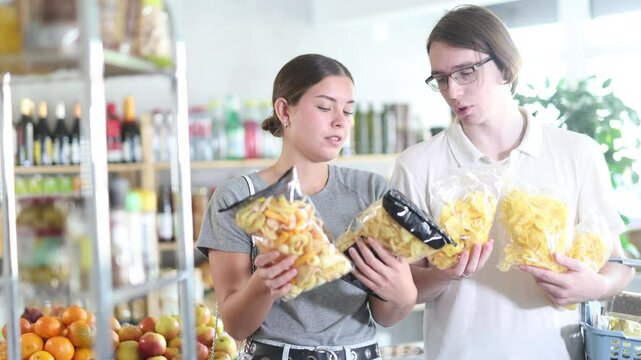 Youth guy with wife choose and buy chips and potato mix crisps in store. Visitors make choice of assortment, buy potato snack crispy rings, sticks. Clients hesitate before buying