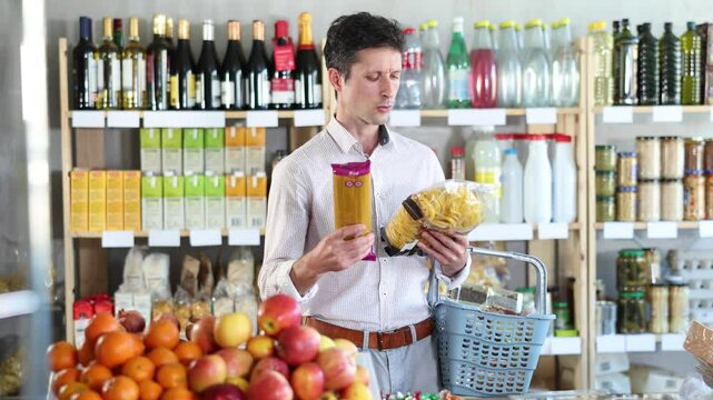 Man in light shirt took some varieties of pasta from shelf. Client compare pasta noodles items package. Customer examines contents of package, choose noodles garnish product