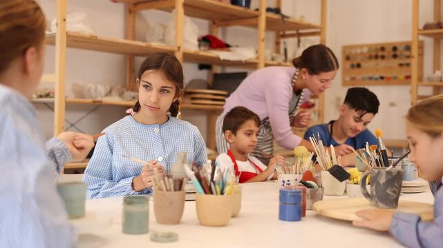  Teenagers and children at a ceramics workshop in an art studio. Woman teacher shows how to paint clay dishes with a brush