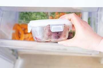 Female hand holding glass box with frozen cherries against refrigerator background.