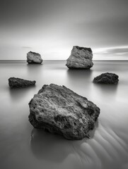 Naklejka premium A serene black and white seascape featuring large rocks emerging from calm waters under a soft, cloudy sky.