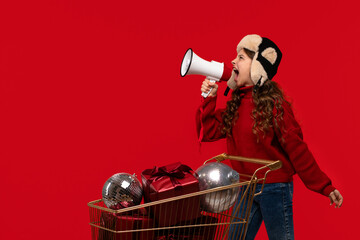 Excited girl in a red sweater and fur hat shouts into a megaphone next to a gold shopping cart full...