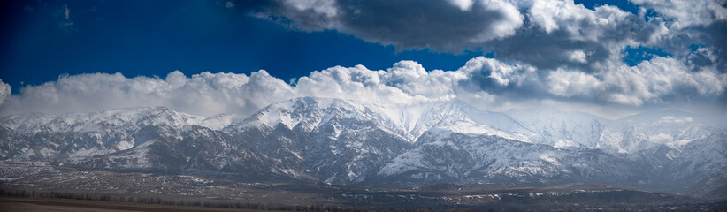 Obraz premium he image shows a majestic mountain range covered in snow under a dramatic sky filled with large, fluffy clouds. The mountains have a rugged and steep appearance, with various peaks and ridges visible.