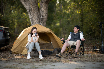 A man and a woman are sitting together in front of a camping tent, The woman is drinking from a mug.