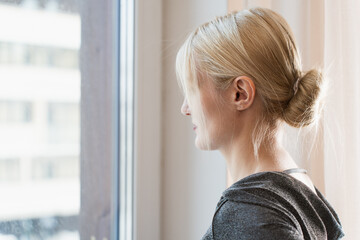 A blonde woman in a stylish outfit gazes out of a window, lost in thought. The soft natural light...