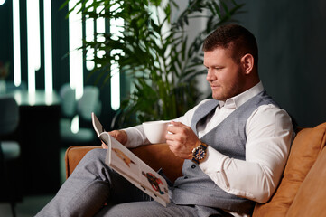 Young man in a suit with a cup in his hands watching a magazine. Businessman during a break sits on...