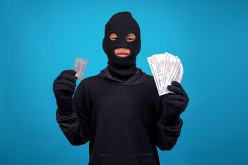 Mysterious Masked Man And Thief Showing Some Stolen Money and Credit Card Against Blue Background