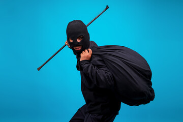Housebreaker In Balaclava Carrying Bag And Holding Crowbar Isolated On Blue Studio Wall