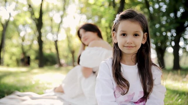 A girl smiles at the camera while sitting in a serene sunlit park, with an adult appearing blurred behind her. The greenery and soft light convey a peaceful and happy atmosphere.
