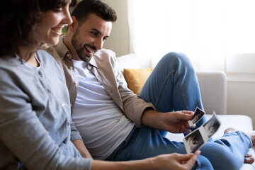 Side view of smiling pregnant couple looking baby ultrasounds at home sitting on the couch.