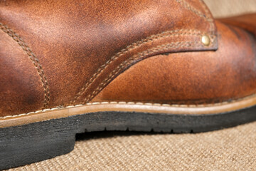 Close-up of a worn brown leather boot with visible creases, patina, and sturdy stitching. The rustic background enhances the rugged, vintage aesthetic.