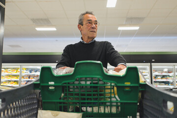 Elderly man pushing a shopping cart filled with groceries in a supermarket, navigating through the...