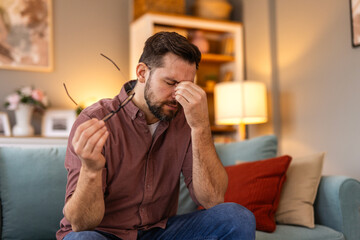 A stressed man sitting on a couch, holding his glasses and rubbing his forehead, overwhelmed with...