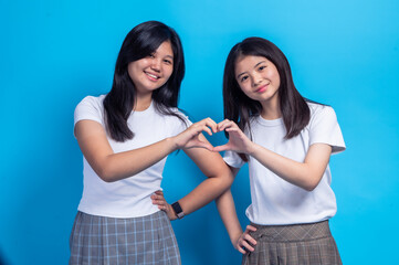 Two Asian teenage girls standing side by side against a bright blue background, smiling and forming a heart shape with their hands, wearing white t-shirts and plaid skirts