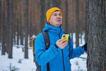 Male hiker dressed in a blue jacket and yellow beanie, using a smartphone gps app to check his location while navigating through a wintery, snowy forest landscape