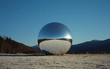 A large, reflective silver sphere sits in a snowy landscape against a clear blue sky. Mountains are visible in the background. The sphere reflects the sun and the surrounding environment