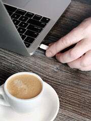 Vertical image of a man plugging in a laptop charging cable near a white cup of espresso coffee.