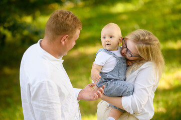 Fototapeta premium In sunny park, mom tenderly holds her precious and sweet baby in her arms, standing next to dad and talking to her toddler, radiating joy and happiness, close up. International Day of Families.