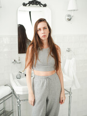 Young and sleepy woman with long hair in pajamas stands against the background of a sink in the...