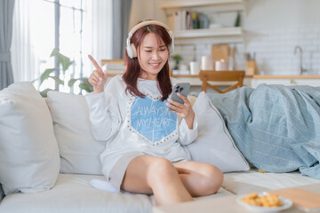 Asian stylish young woman is sitting on a sofa and listening to music with a wireless bluetooth...
