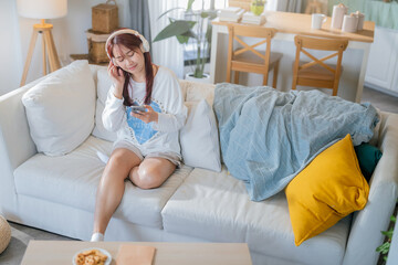 Asian stylish young woman is sitting on a sofa and listening to music with a wireless bluetooth...