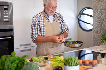 Smiling senior man with beard is busy cooking and holding a pan with vegetables. Elderly person in...