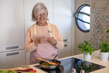 Smiling senior gray haired woman at home cooking vegetables adds some herbs in the pan. People in...