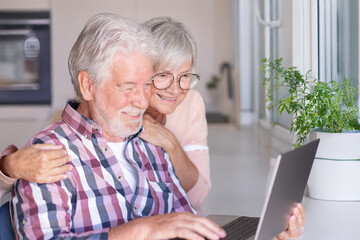 Mature white haired senior husband and wife sitting at home sharing internet connection using...