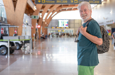 Handsome carefree senior man travels at airport with backpack walking towards board gate. Portrait of elderly happy traveler enjoying trip and vacation