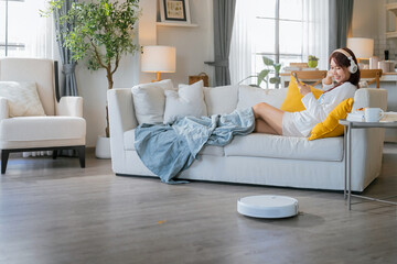 Young Asian woman relaxing sit on sofa home and turning on her smart vacuum cleaner using smartphone control robot while listening music headphone smart life smart technology lifestyle home hygiene
