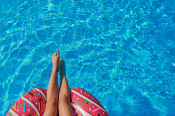 Female legs bask in sunlight near turquoise swimming pool, pink inflatable ring floating lazily. Summer relaxation theme dominates with vibrant colors, summer relaxation ideal for travel blogs