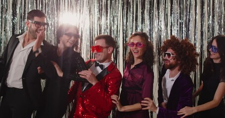 Group of joyful stylish friends standing against sparkling background, doing wave with their hands. People wearing trendy outfits and funky glasses dancing in coordinated moves at retro themed party.