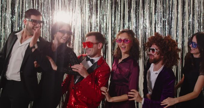 Group of joyful stylish friends standing against sparkling background, doing wave with their hands. People wearing trendy outfits and funky glasses dancing in coordinated moves at retro themed party.