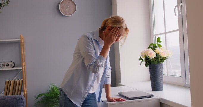 Tired overworked woman feeling dizzy and suffering from terrible headache, going to loose consciousness, posing in living room near window, being exhausted after working at laptop for long time