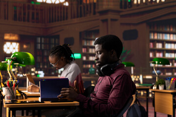 African american male student opening his textbook to complete his handwritten class notes with information from specialty books at the university library. Young adult reading academic papers.