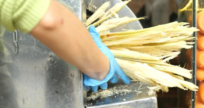 Street vendor crushing sugarcane with manual press, wearing blue gloves and extracting fresh juice amid colorful market atmosphere, showcasing local street food preparation