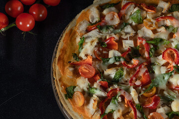 Rustic homemade pizza featuring roasted chicken, colorful bell peppers, cherry tomatoes, fresh basil, melted mozzarella on dark backdrop with additional tomatoes nearby