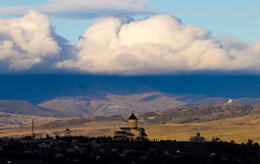 A picturesque Makhata Iveron Icon Church sits nestled in the hills as dramatic clouds cast shadows over the mountainous landscape at sunset in Georgia, creating a beautiful contrast.